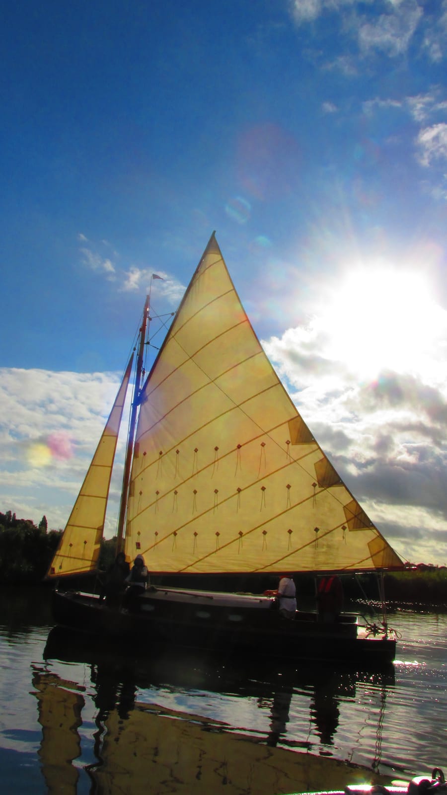A wooden sailing boat under a tall sail, backlit by low evening sun on calm water